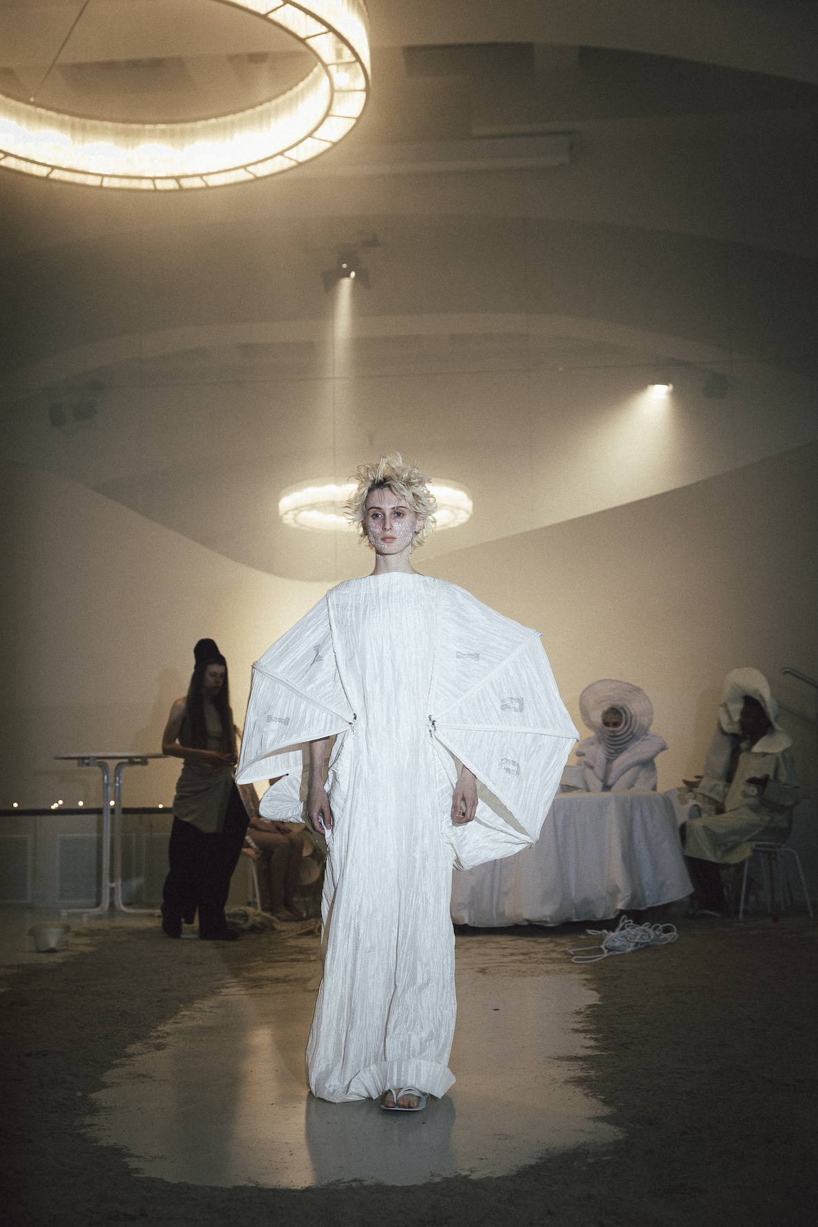 A performer in a long white garment stands beneath a circular ceiling light during ANTICON.